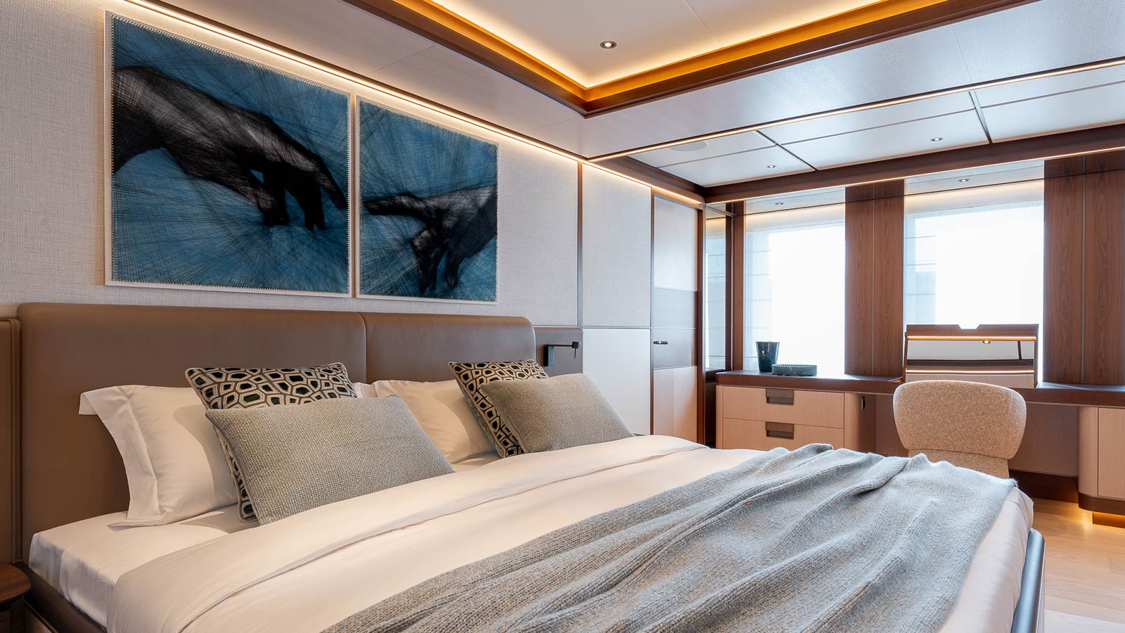 A modern, elegant bedroom with a double bed featuring decorative pillows and a soft gray throw blanket. Above the bed hangs a diptych artwork depicting hand gestures on a blue background. The room is well-lit with a sleek wooden wardrobe and a desk by the window, accompanied by a cream-colored chair.
