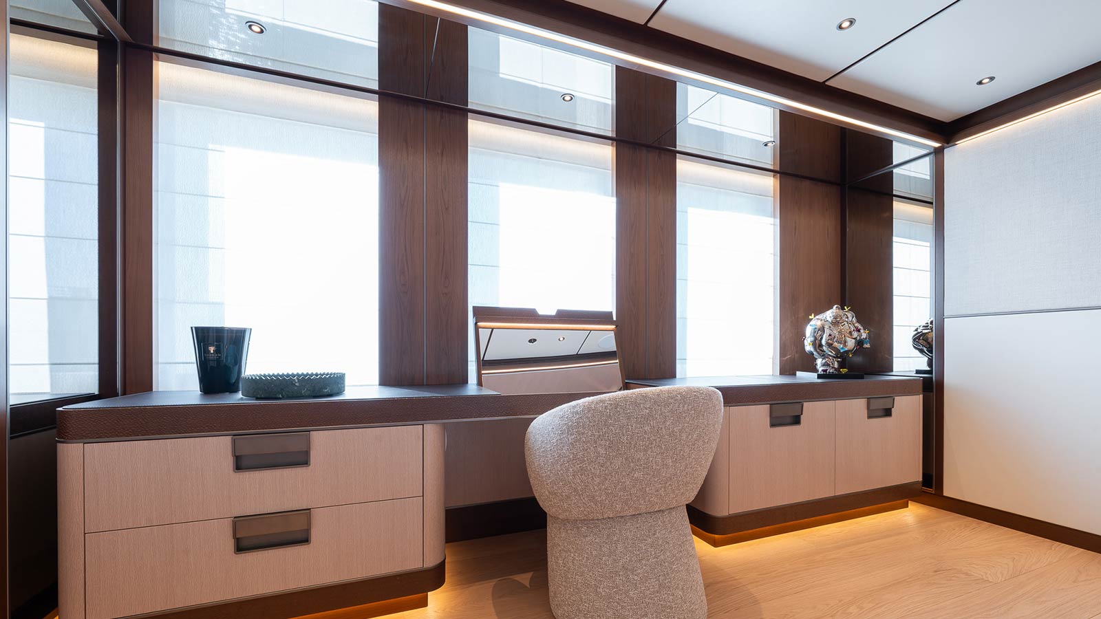 A modern interior space with a light wood desk, a padded chair, decorative objects, and large windows letting in natural light.