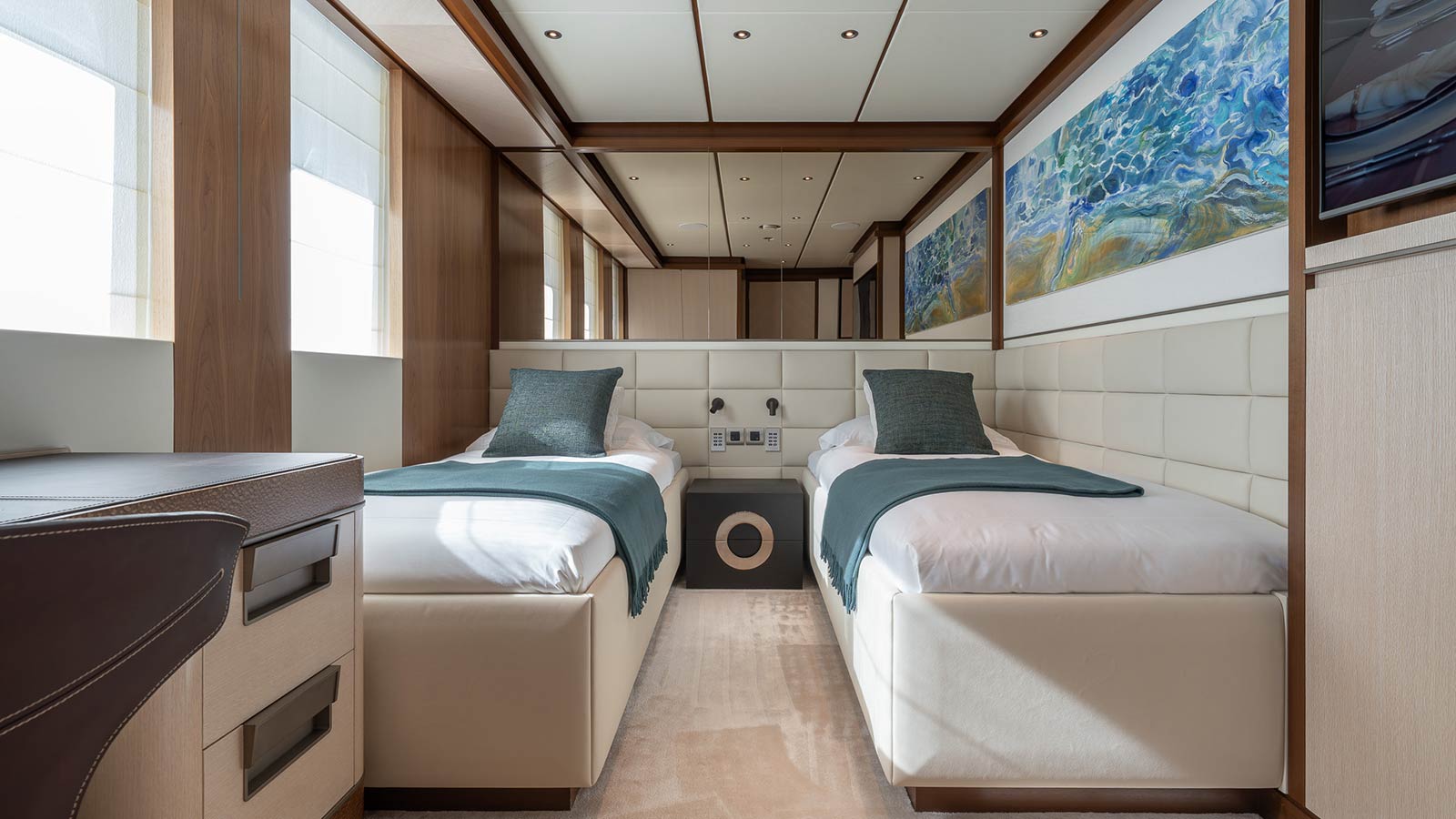 A luxurious cabin featuring two single beds with white bedding and teal blankets and pillows, separated by a small nightstand. The room has wood paneling, soft lighting, and large windows on the left side. A vibrant abstract painting decorates the wall above the beds.