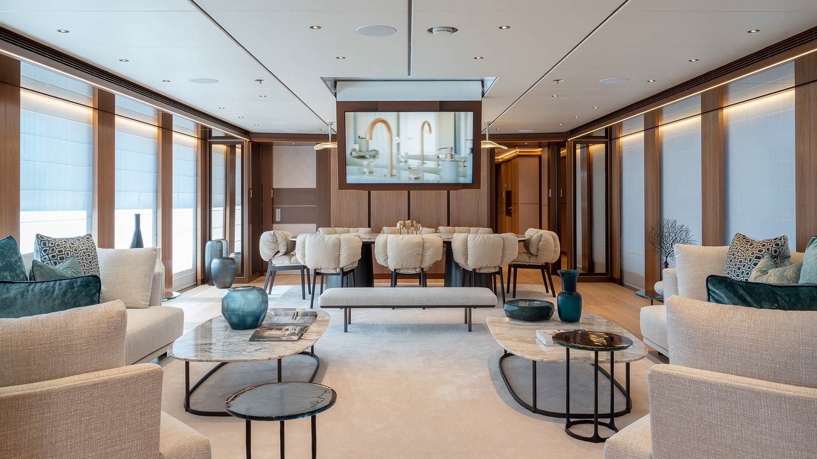 Luxurious yacht interior featuring a spacious living area with plush beige sofas, decorative cushions, marble coffee tables, and a large screen on the wall. The room is accentuated by wooden panels, elegant decor pieces, and ample natural light from large windows.