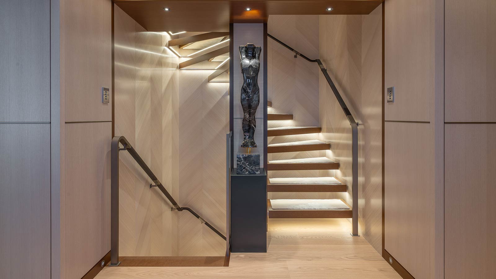 A modern, well-lit stairwell with wooden walls and an illuminated staircase, featuring a black marble sculpture on a pedestal at the center.