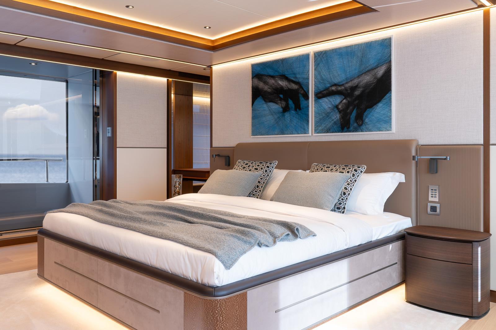 A modern bedroom with a large bed covered in white linens and a light blue blanket, two patterned pillows, wooden side table, and two abstract hand paintings on the wall. The room features warm, ambient lighting and large windows with a view of the sea.