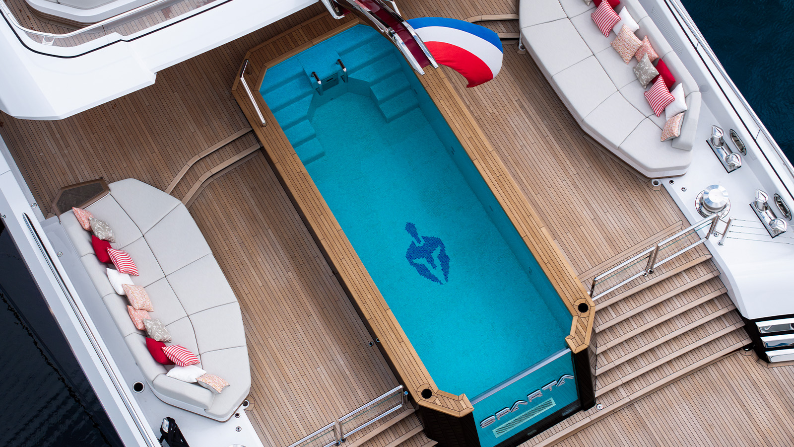 Aerial view of a luxury yacht's deck featuring a rectangular pool with steps on one end and a decorative Spartan helmet design at the bottom. The deck has wooden flooring with seating areas on either side adorned with cushions in red, white, and beige. A set of stairs leads to the lower deck, and a flag with blue, white, and red stripes is visible.