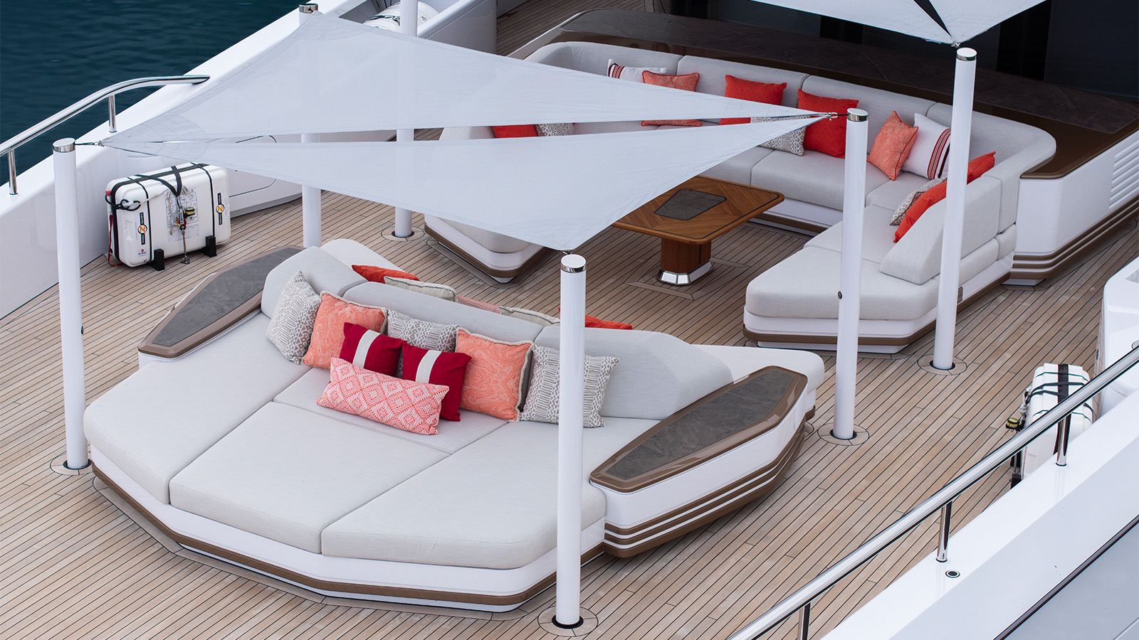 A luxurious yacht deck featuring modern, white cushioned seating areas with colorful accent pillows and a triangular sunshade canopy above. The deck is made of wood planks, adding to the elegant design, and includes two white equipment cases near the railing.
