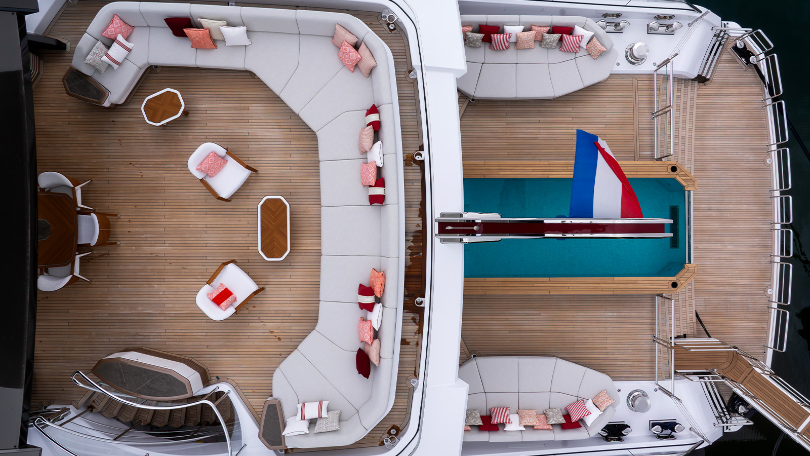 Aerial view of a yacht's deck featuring a large seating area with white cushioned sofas adorned with colorful pillows. Two small wooden tables are placed between the sofas and there are additional chairs with pillows. Adjacent to the seating area is a pool with a red and blue flag, surrounded by teak decking.