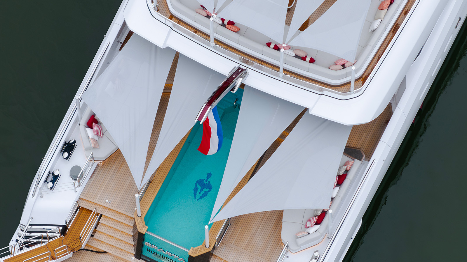 Aerial view of a luxury yacht's deck featuring a swimming pool with a blue design, red and white striped cushions, and spacious seating areas. The yacht is docked near the water, and a flag is displayed at the rear.