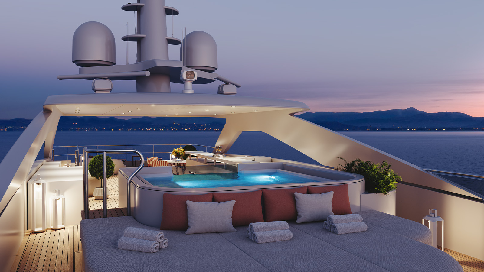 Luxurious yacht deck featuring a jacuzzi, plush seating with cushions and towels, ambient lighting, and views of the ocean at twilight with distant mountains on the horizon.