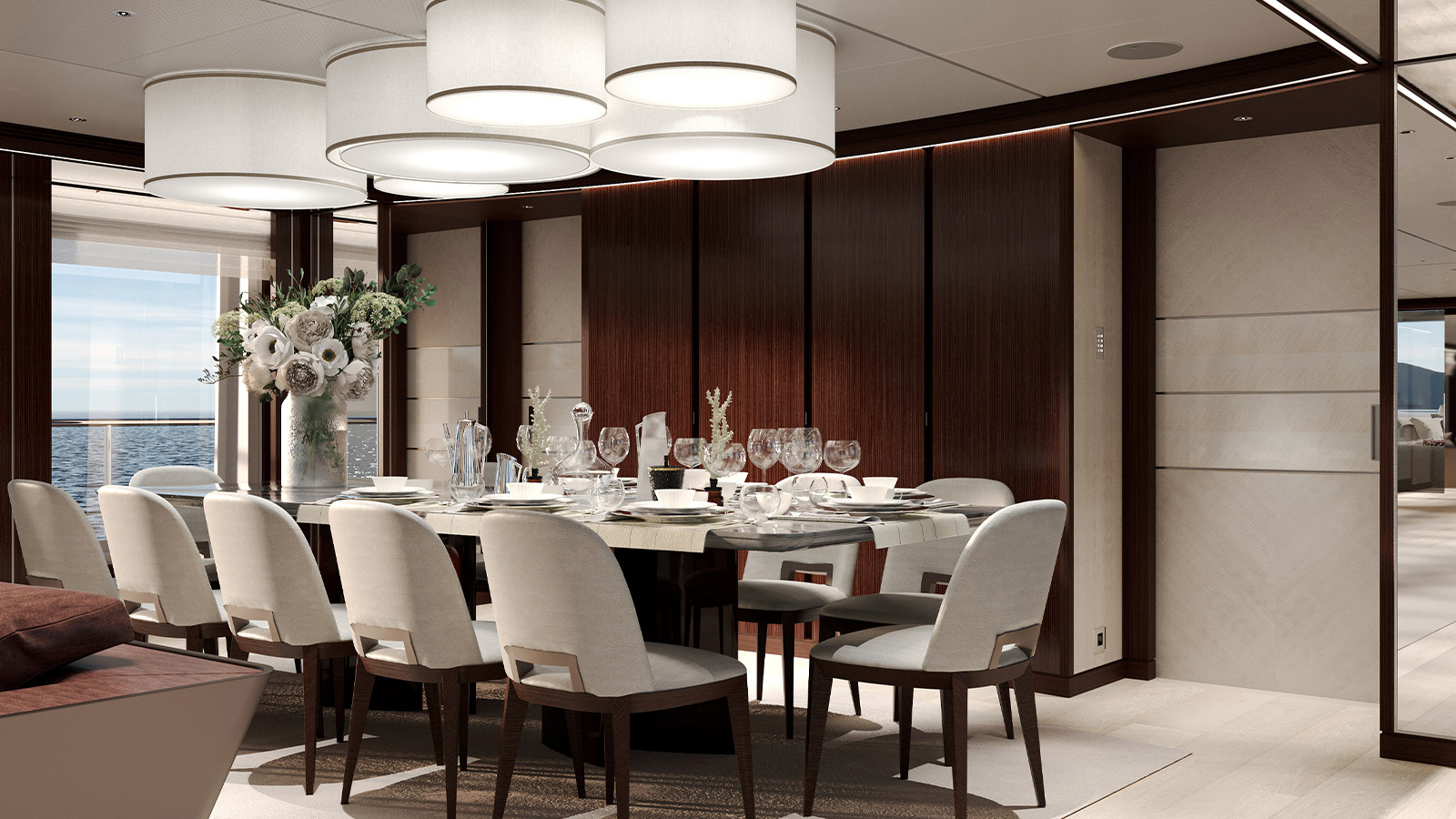 Elegant dining room on a yacht with a large rectangular table set for dining, surrounded by cushioned chairs. The room features modern lighting fixtures above and a vase of flowers as a centerpiece, with ocean views visible through large windows.