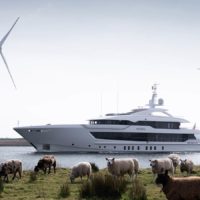 A large yacht sailing on a river with sheep grazing on the grassy riverbank in the foreground and wind turbines visible in the background.
