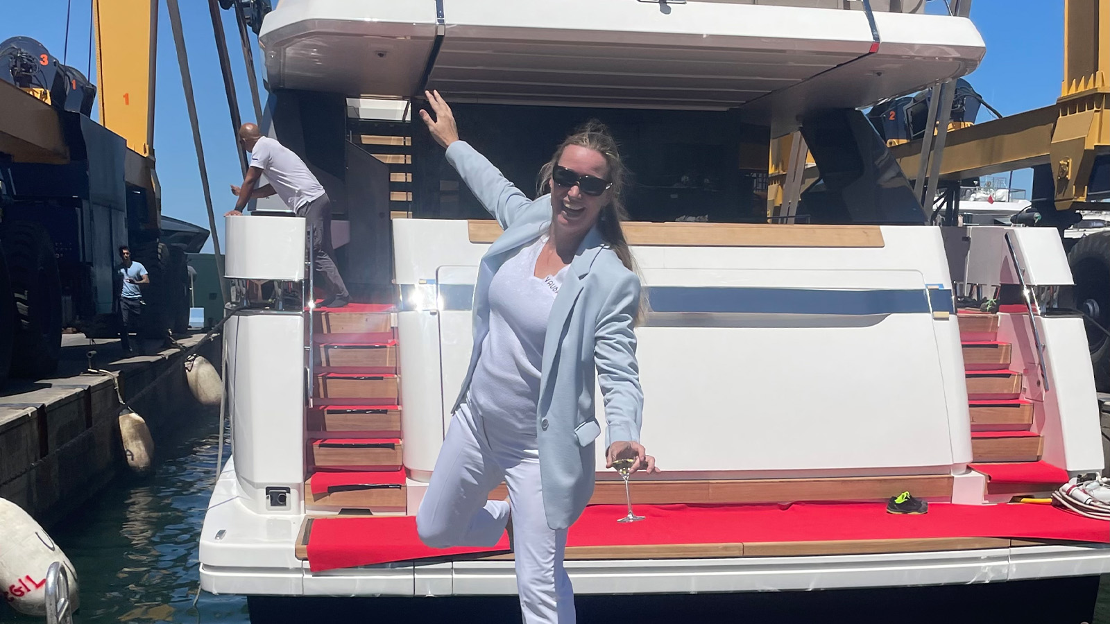 A person in a light blue blazer and white outfit poses on the back of a yacht, holding a wine glass, with a man working nearby on the dock.