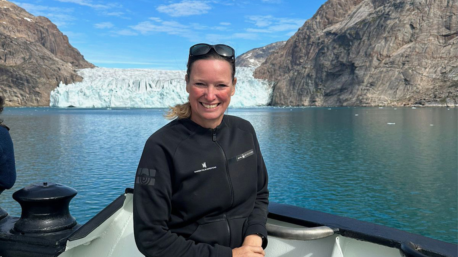 A person smiling and standing on a boat with a glacier and mountains in the background under a clear blue sky.