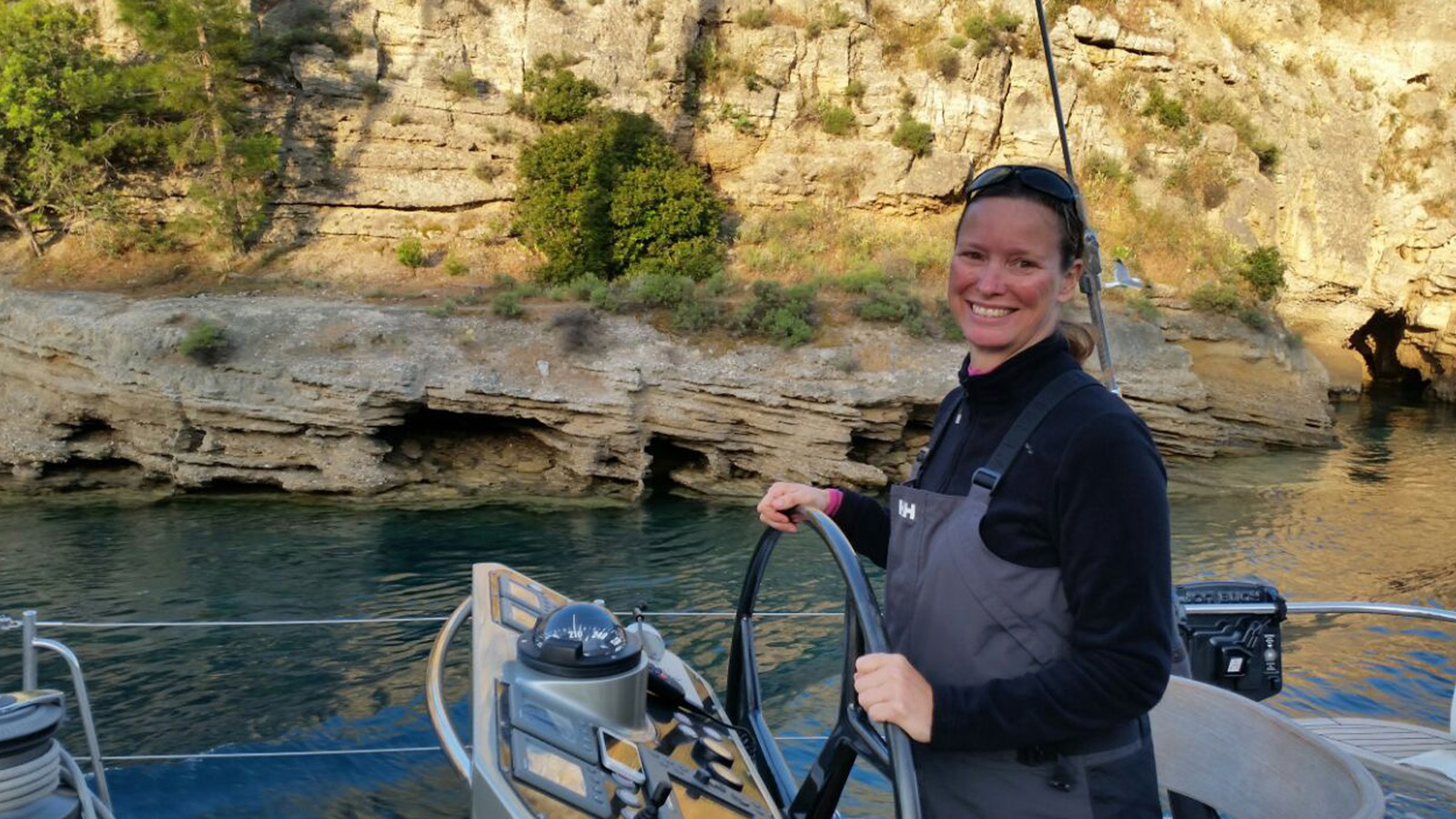 A woman smiling and steering a sailboat, with a rocky coastal landscape in the background.
