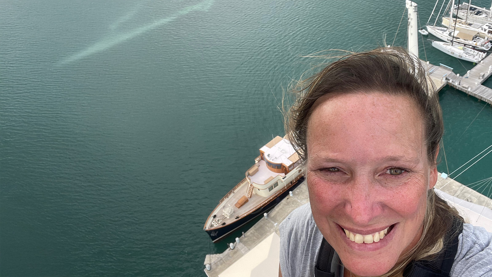 A person smiling at the camera while standing high above a body of water, with a large boat and dock visible below.