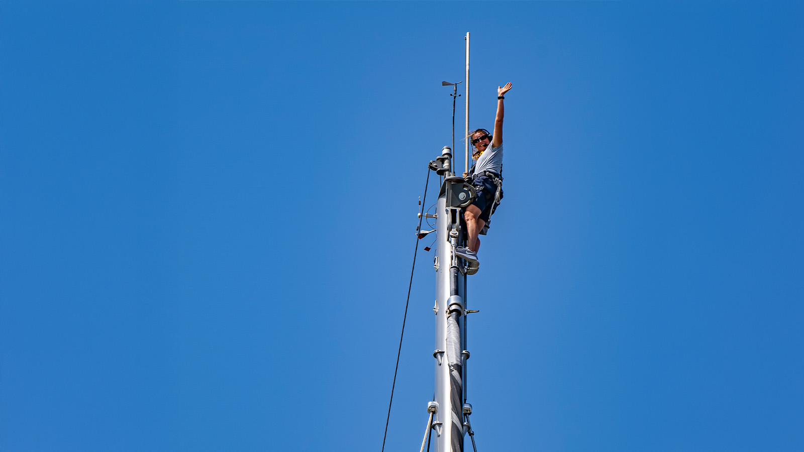 A person in climbing gear waves from the top of a tall mast against a clear blue sky.