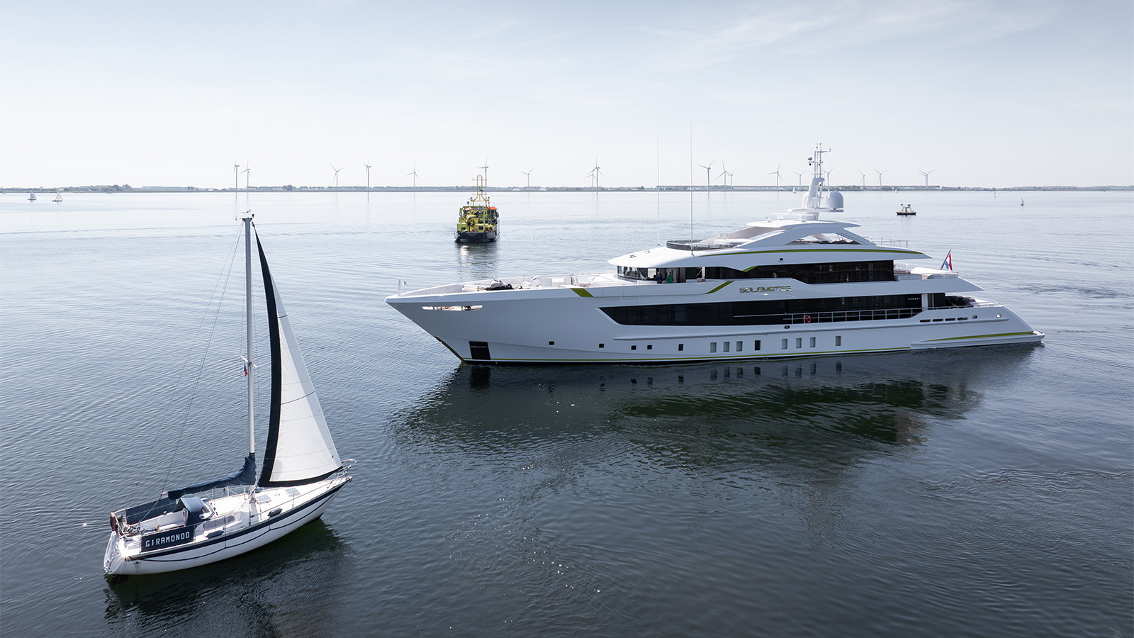A large, modern yacht and a small sailboat named Giramondo on calm water, with wind turbines visible in the distance.