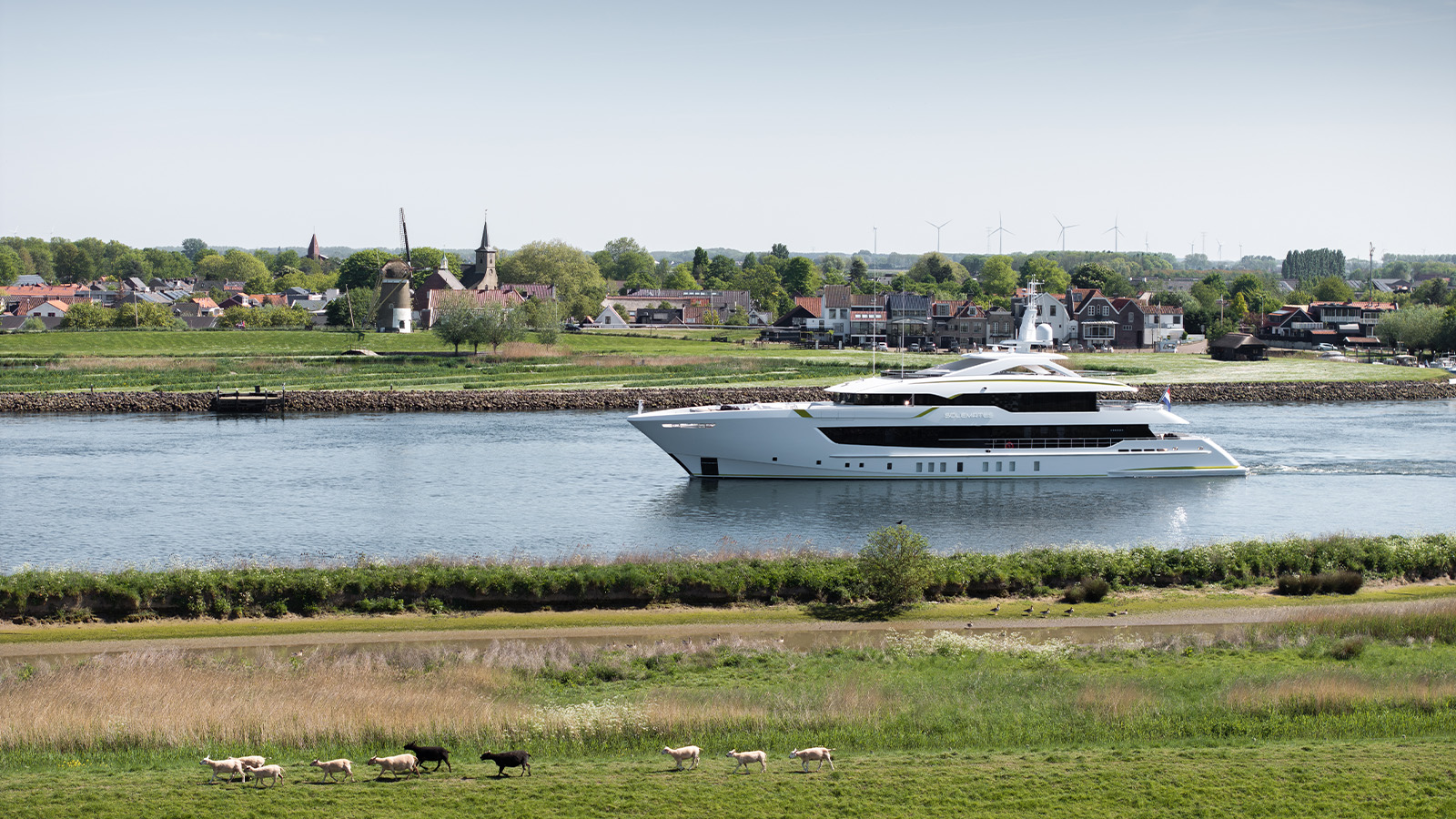 A luxury yacht sailing on a river with a picturesque town and windmill in the background; sheep graze on grass in the foreground.