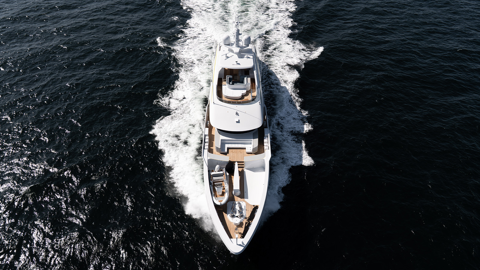 A sleek white yacht cruising through dark blue water, viewed from above, leaving a trail of white wake behind it.