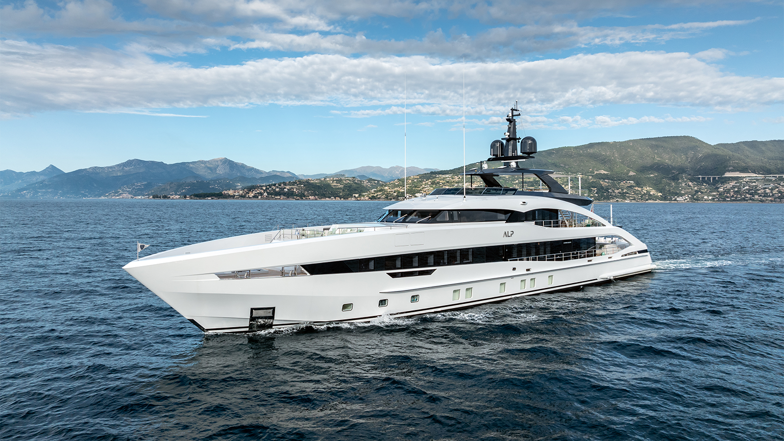 A large, sleek white yacht cruising on the open sea with a mountainous coastline in the background.