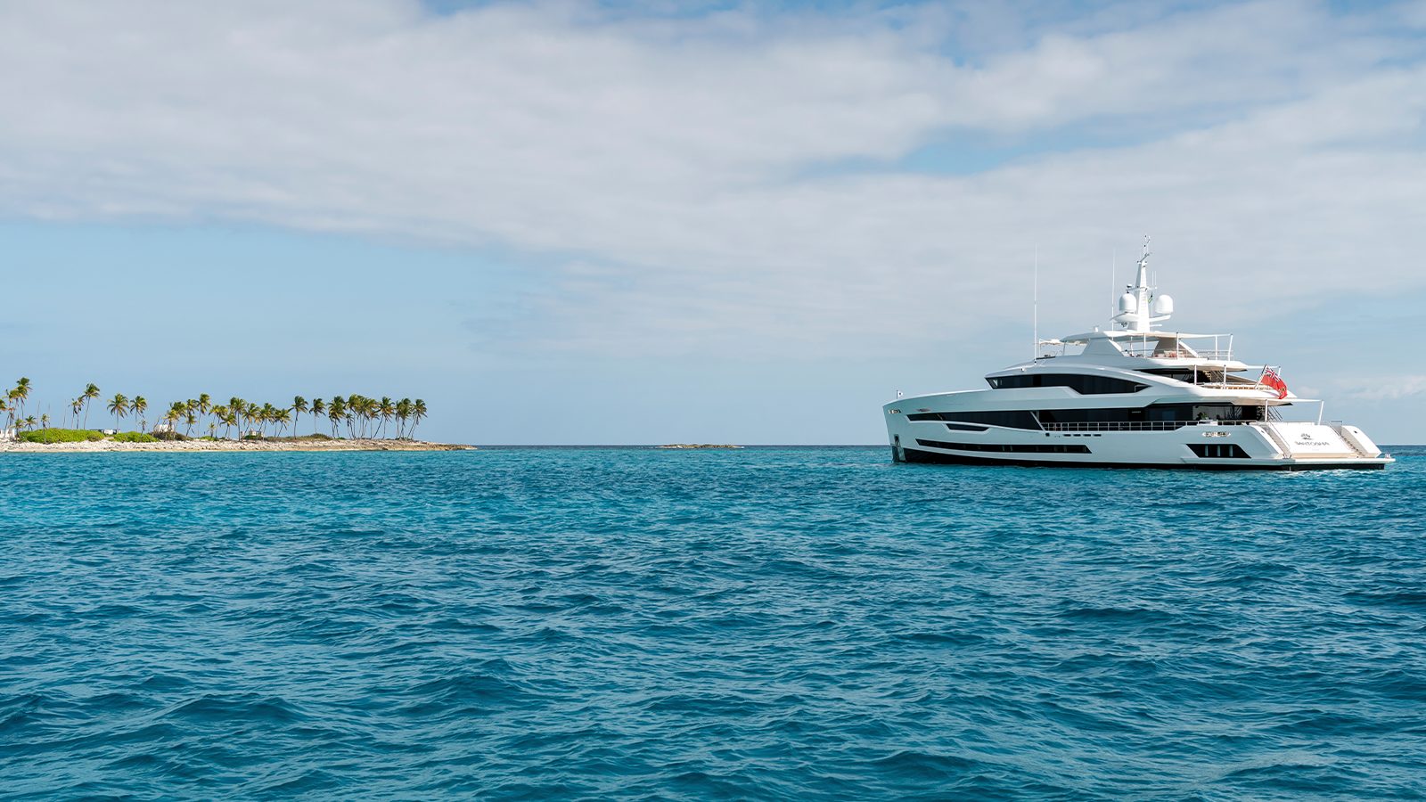 A luxury yacht anchored in a clear blue ocean near a small tropical island with palm trees under a partly cloudy sky.