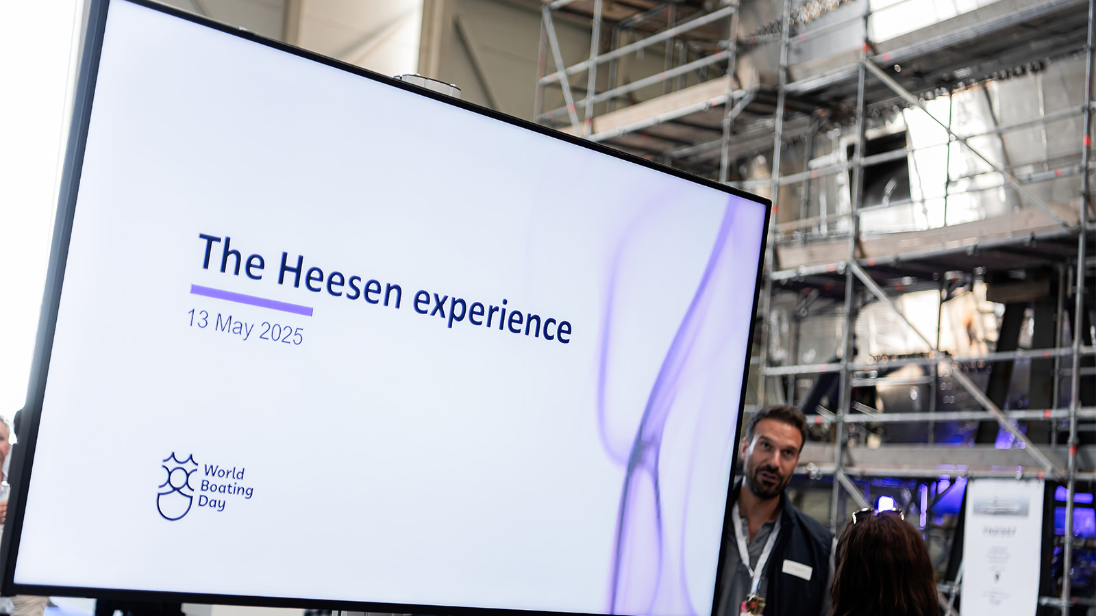 A presentation screen displaying "The Heesen experience 13 May 2025," with a logo and the text "World Boating Day" at the bottom left, set against a background with scaffolding and a person standing nearby.