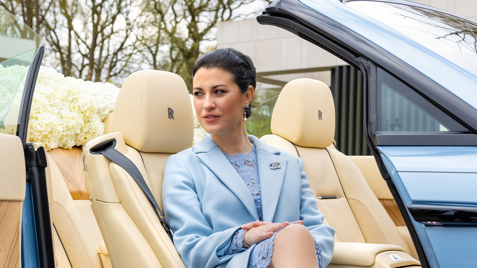 A woman in a blue outfit sits in the back seat of a luxury convertible car with a cream interior, branded with the Rolls-Royce logo, surrounded by a floral arrangement.