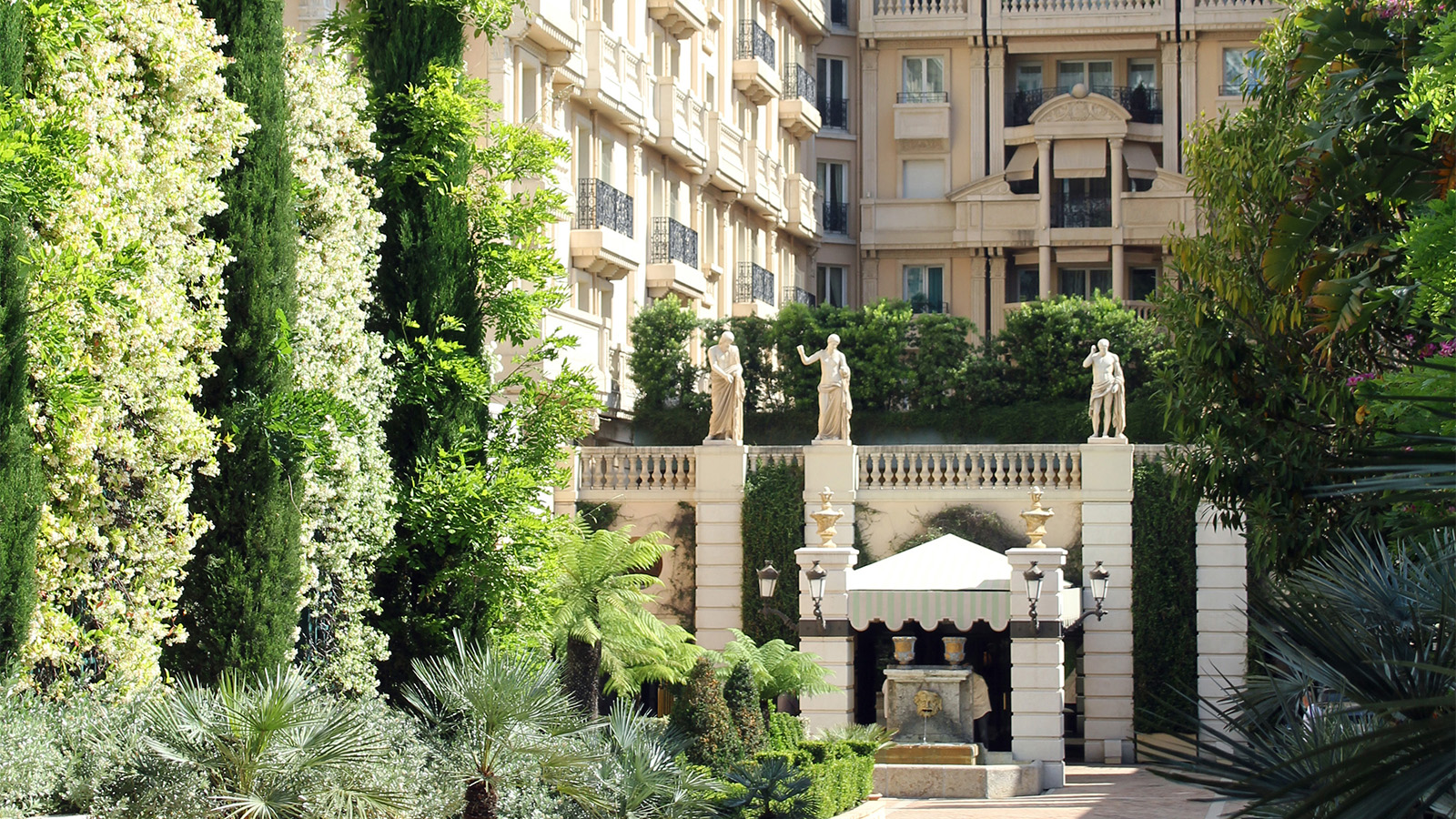 A luxurious building surrounded by lush greenery and classical sculptures; columns supporting statues are situated at the entrance.