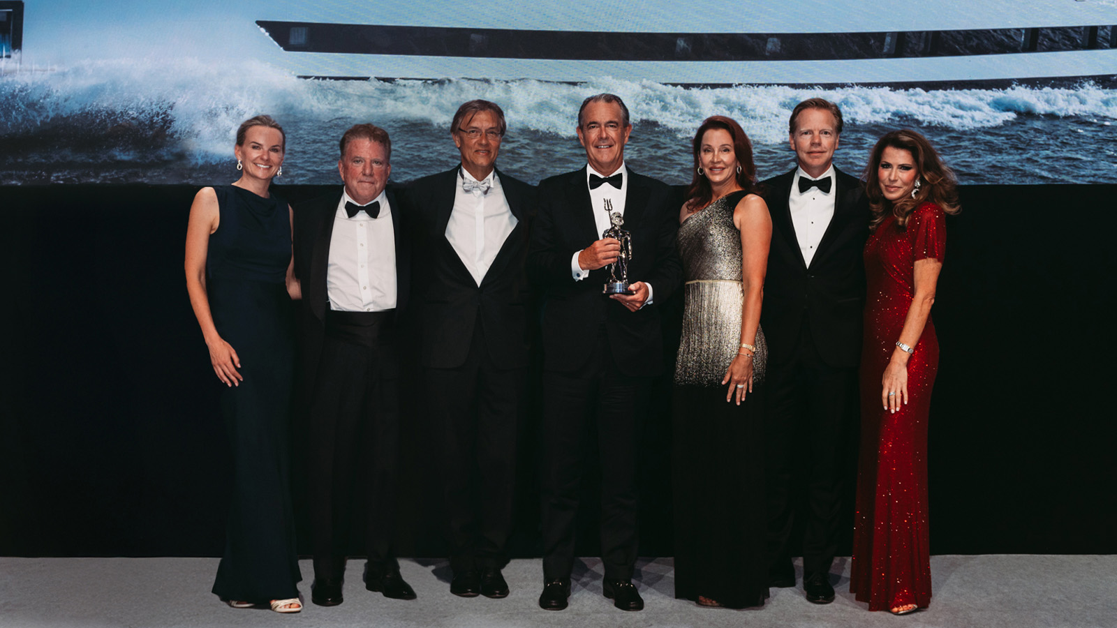 A group of six formally dressed individuals, three men in tuxedos and three women in elegant evening gowns, posing together; one man is holding a trophy. A large screen behind them displays an image of a yacht cutting through waves.