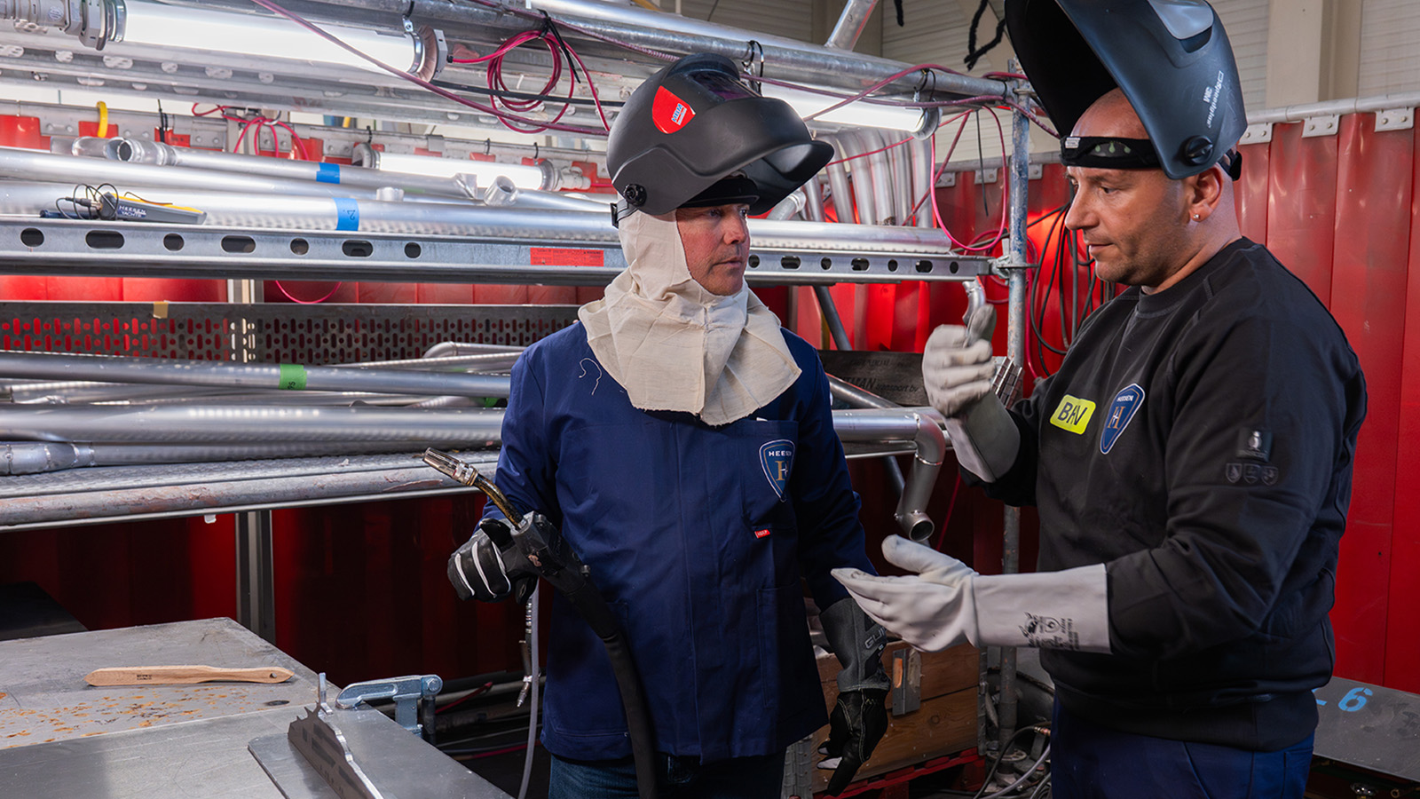 Two workers wearing protective gear, including helmets and gloves, are in a metalwork setting, with one holding welding equipment and the other gesturing as they discuss a task.