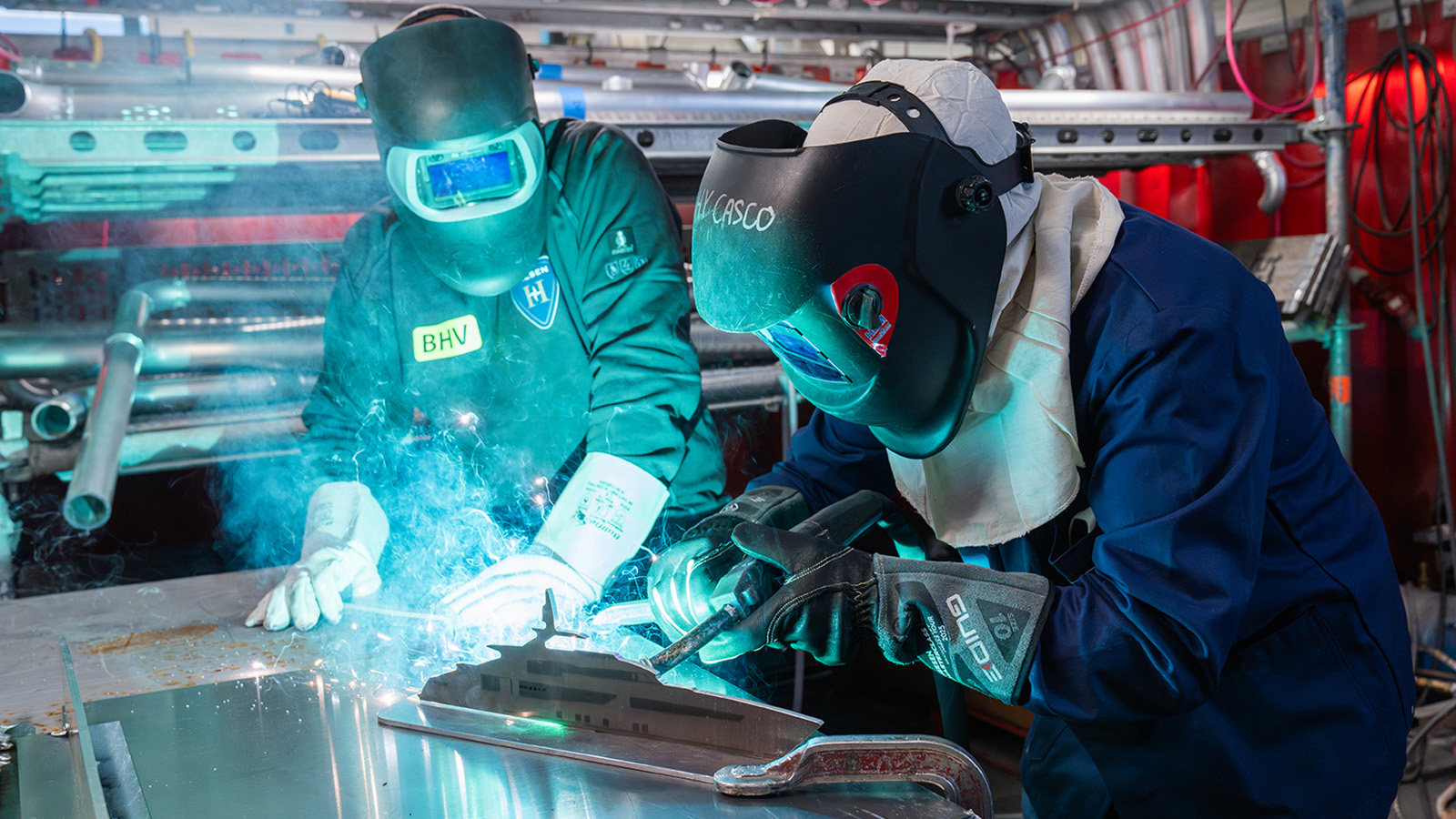 Two people wearing protective welding helmets and gloves work on a metal surface in a workshop, with sparks and bright light from welding visible.