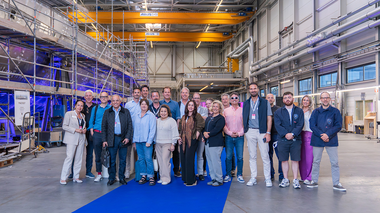 A group of people posing for a photo in an industrial setting, with scaffolding and equipment visible in the background. They are standing on a blue carpet, suggesting a formal event or tour.