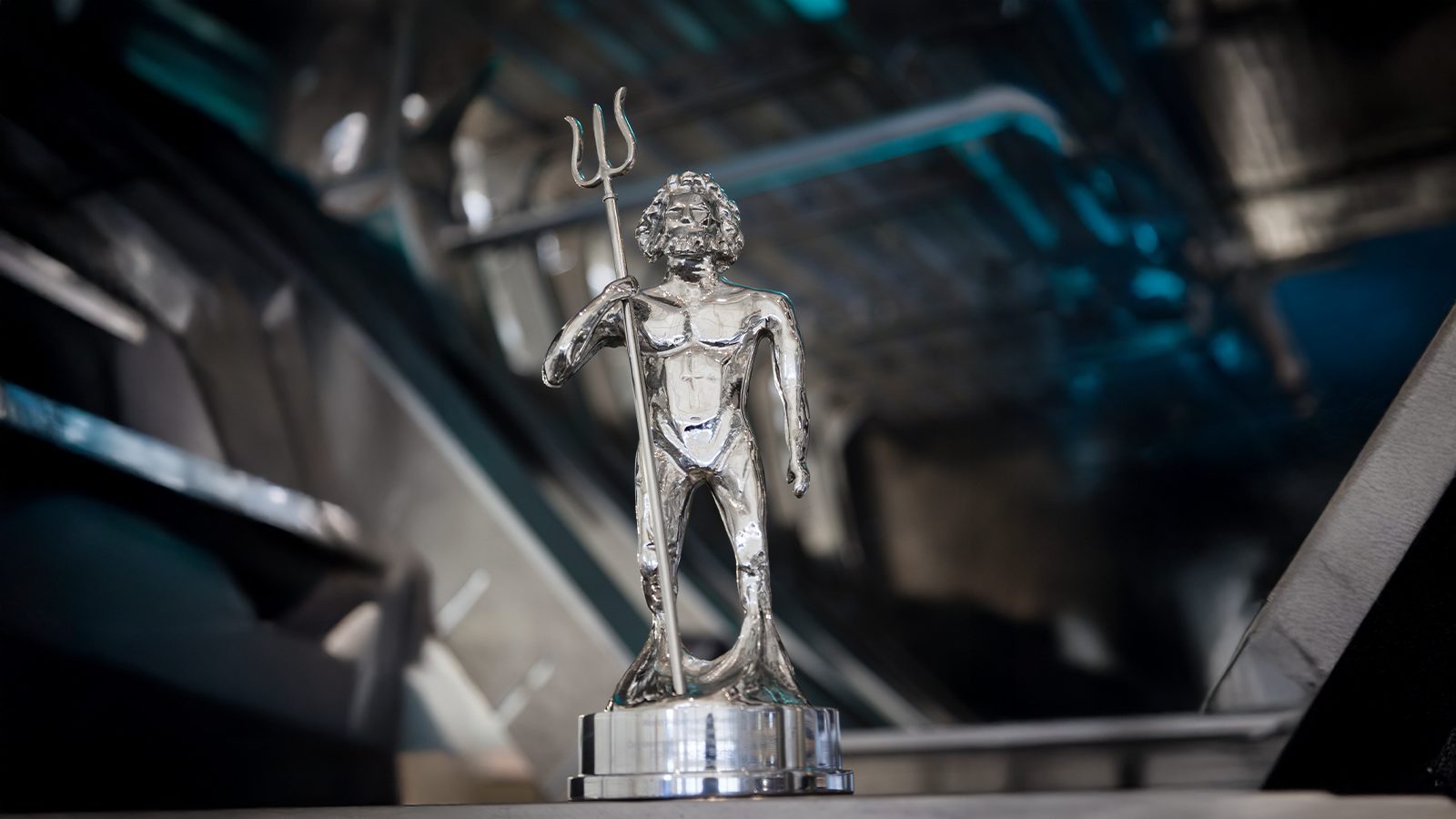 A silver trophy depicting a stylized figure of a man holding a trident, standing on a reflective base. The design is minimalist with an emphasis on smooth, flowing lines. The background is blurred, featuring metallic and blue tones.