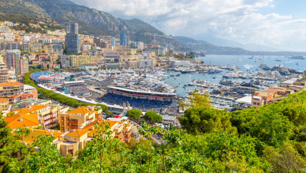 A panoramic view of Monaco, featuring a marina filled with yachts, grandstands, and a cityscape with colorful buildings and lush greenery in the foreground. Hills rise in the background under a partly cloudy sky.