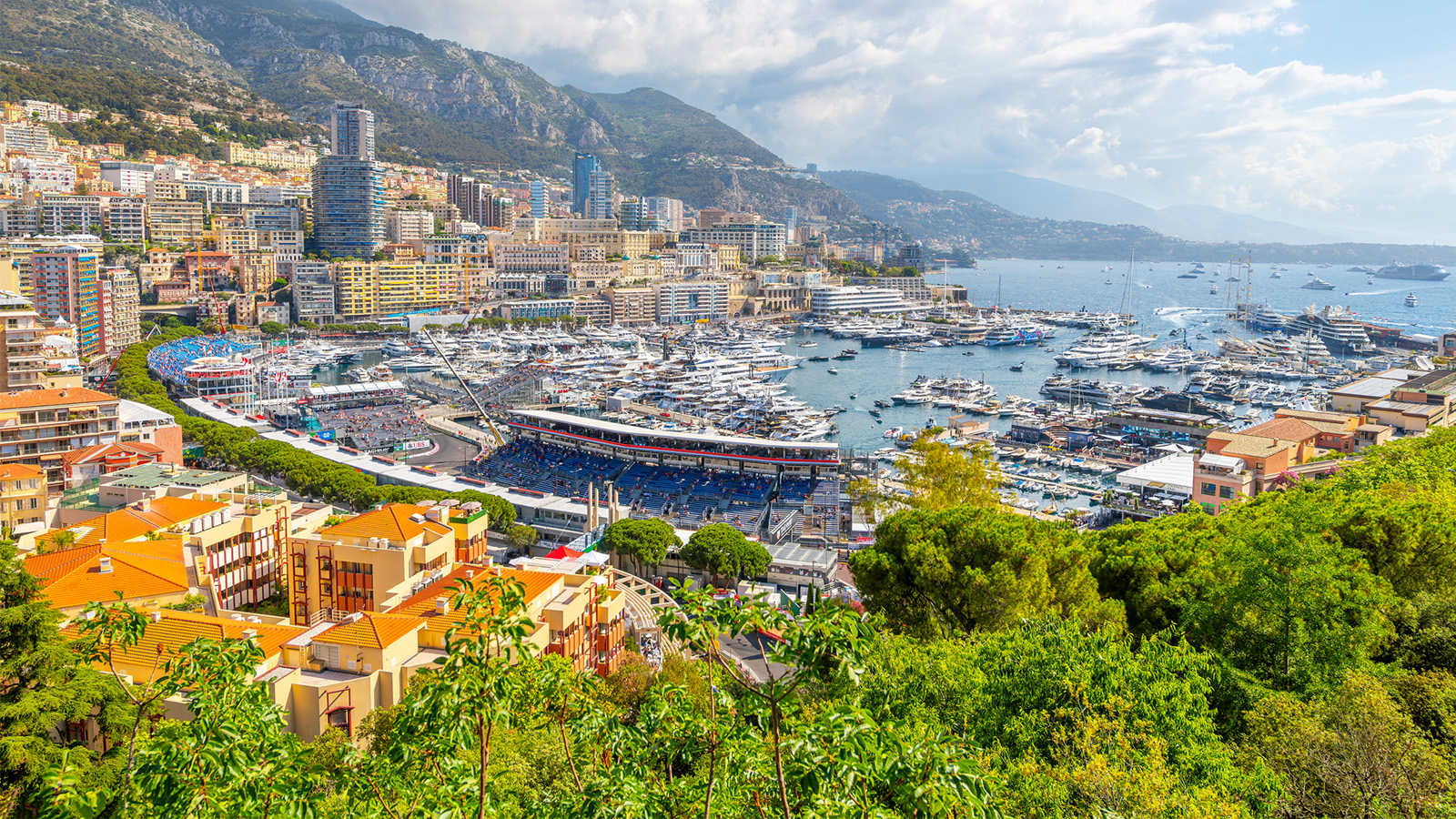 A panoramic view of Monaco, featuring a marina filled with yachts, grandstands, and a cityscape with colorful buildings and lush greenery in the foreground. Hills rise in the background under a partly cloudy sky.