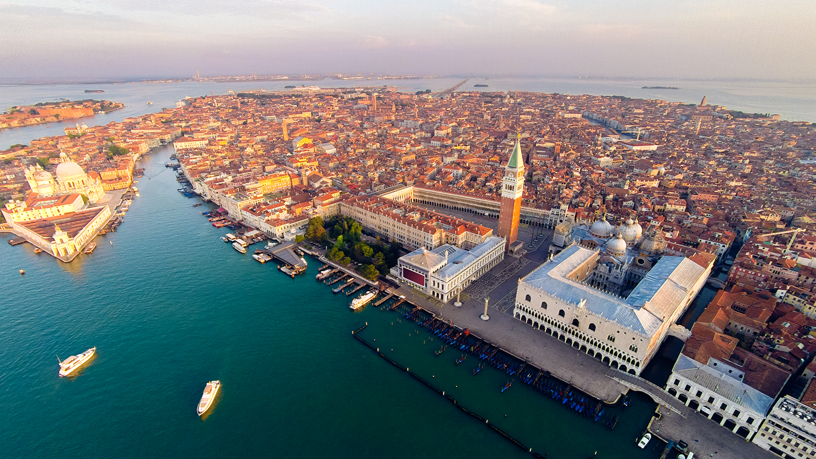 Aerial view of Venice, Italy, showcasing St. Mark's Square with its prominent campanile, the Doge's Palace, and surrounding buildings. The Grand Canal and several other waterways weave through the densely packed cityscape, with numerous boats navigating the turquoise waters.