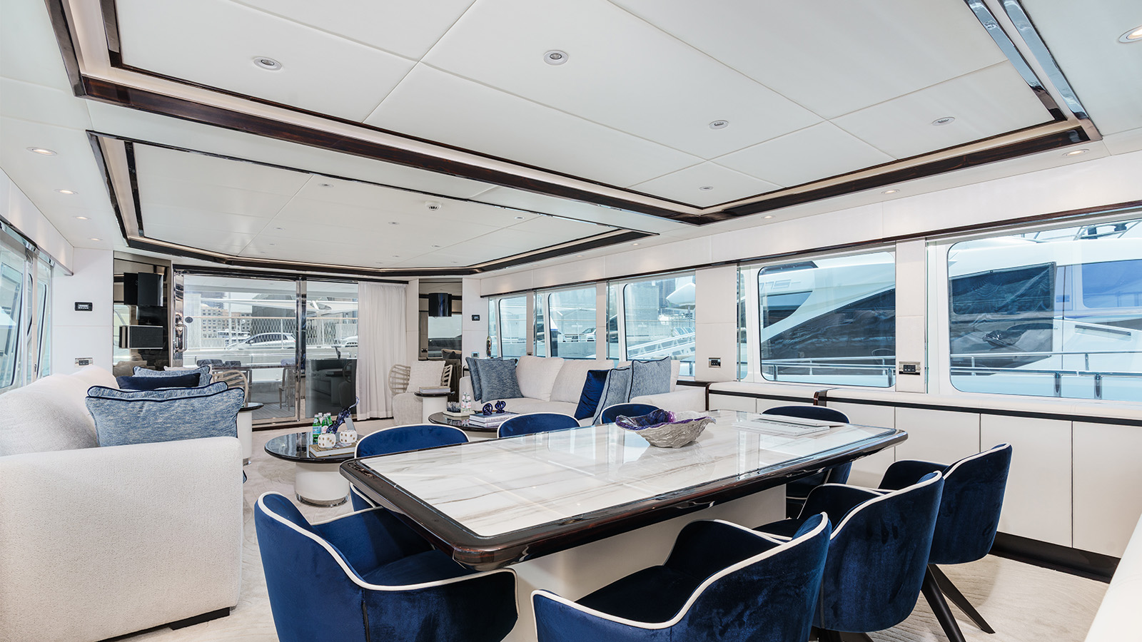 Luxurious interior of a yacht featuring a spacious dining area with a large, marble-topped table and blue velvet chairs, adjacent to a comfortable seating area with sofas and a view of marina through large windows.