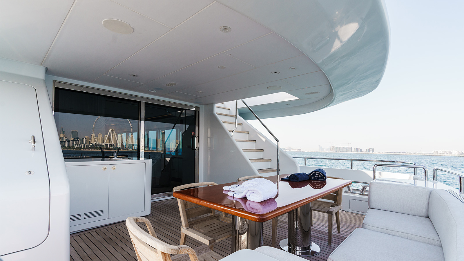 Spacious yacht deck with a wooden table, chairs, and a staircase leading to an upper level, overlooking a city skyline and Ferris wheel.