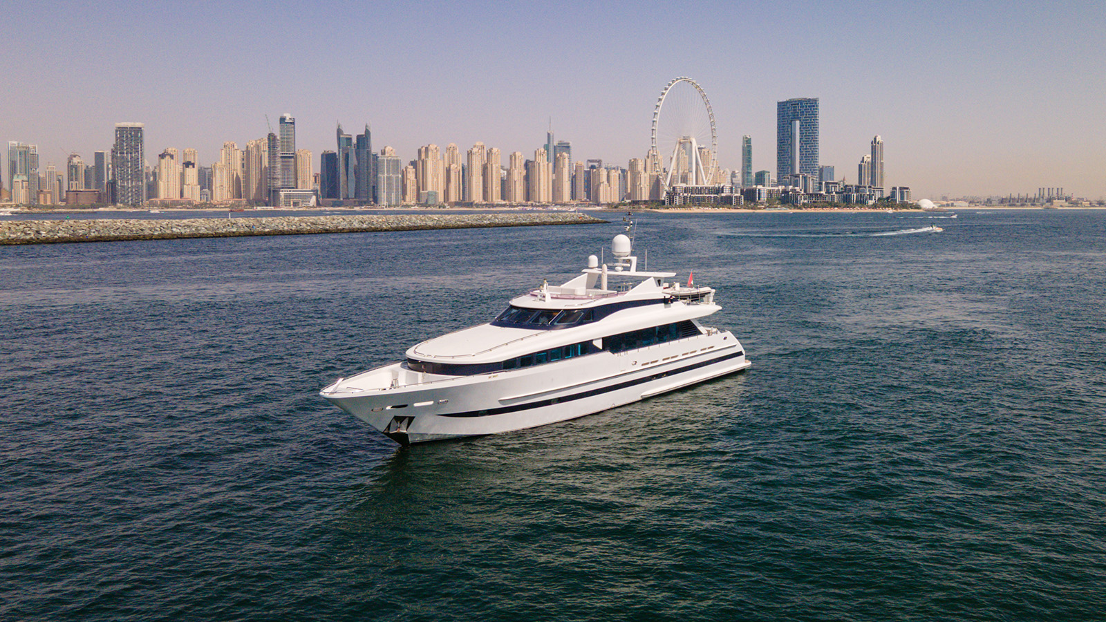 A large white yacht sailing on a body of water with a city skyline featuring skyscrapers and a Ferris wheel in the background under a clear sky.