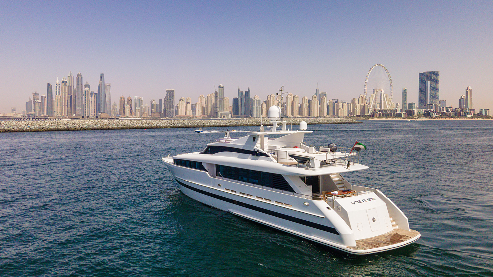 A luxury yacht sails in front of a city skyline with modern skyscrapers and a large Ferris wheel, set against a clear blue sky.