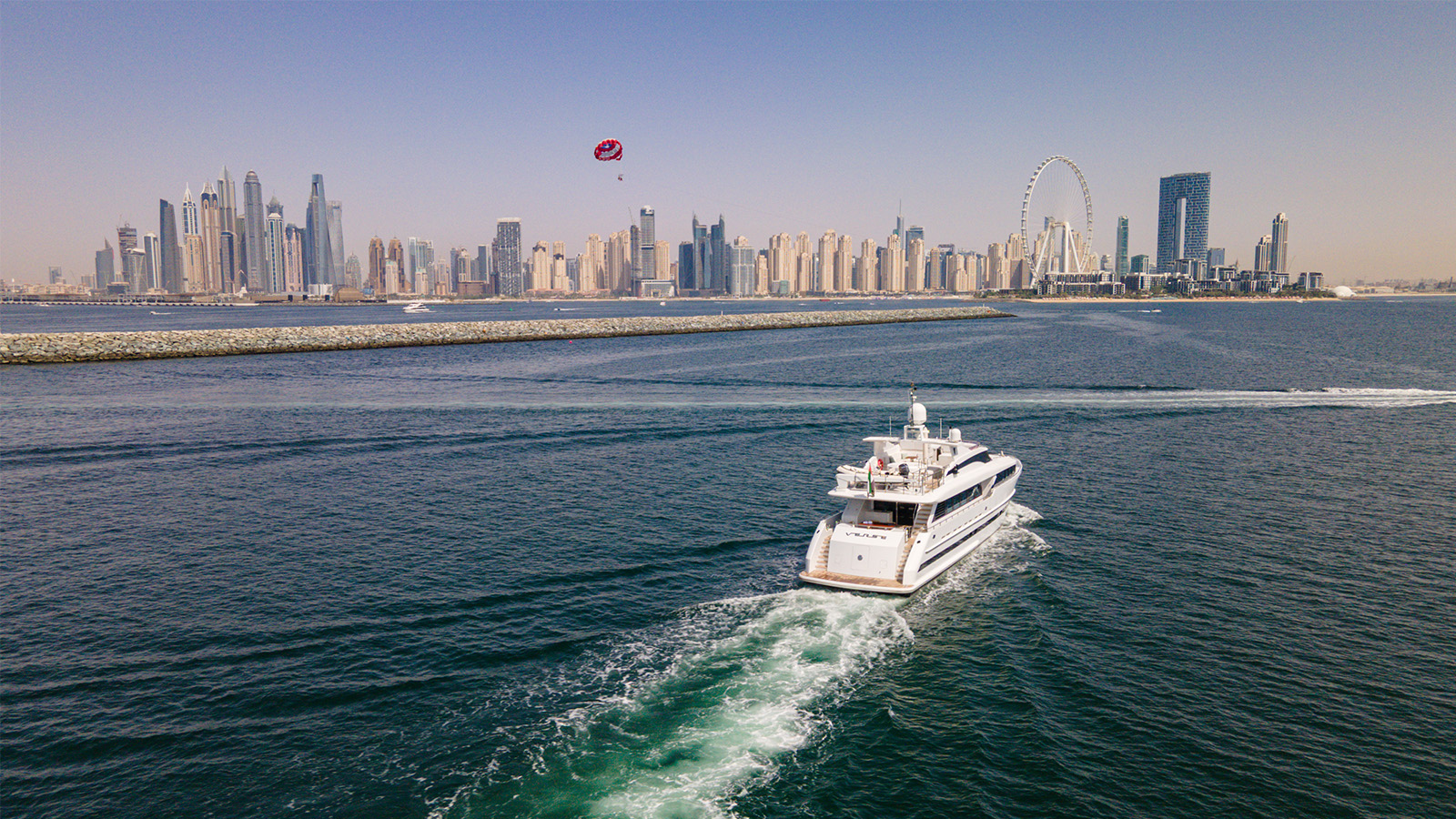 A yacht sailing in the Arabian Gulf with the Dubai skyline in the background, featuring tall skyscrapers and the Ain Dubai Ferris wheel under a clear blue sky.