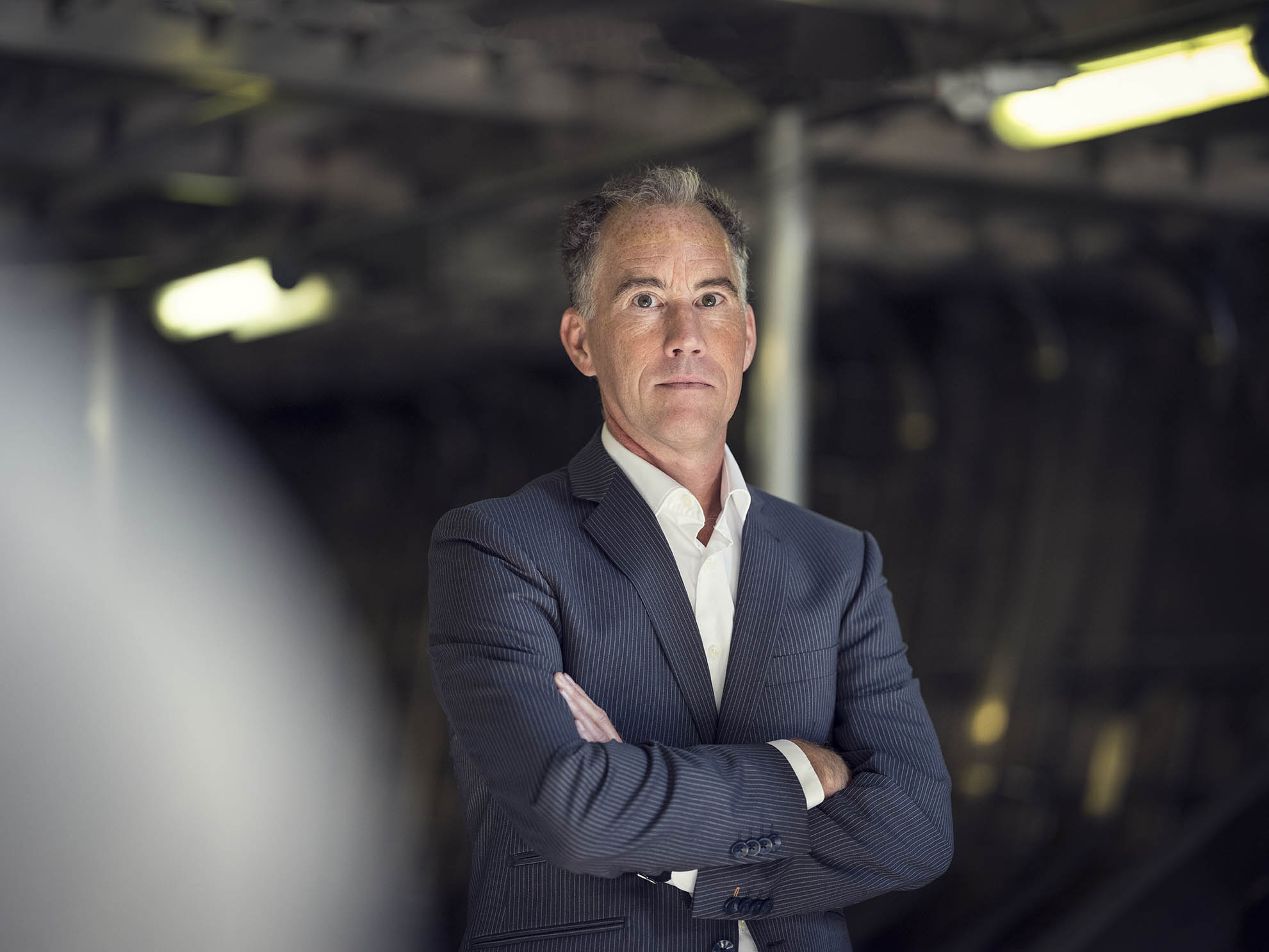 A person in a suit with arms crossed, standing indoors with a blurred industrial background.