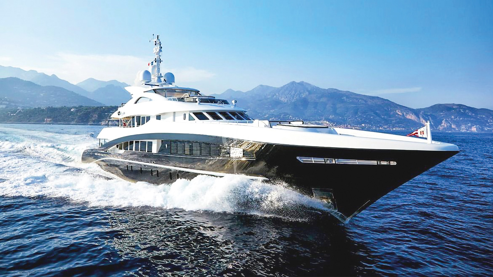A luxury yacht cruising on blue waters with mountainous coastline in the background.