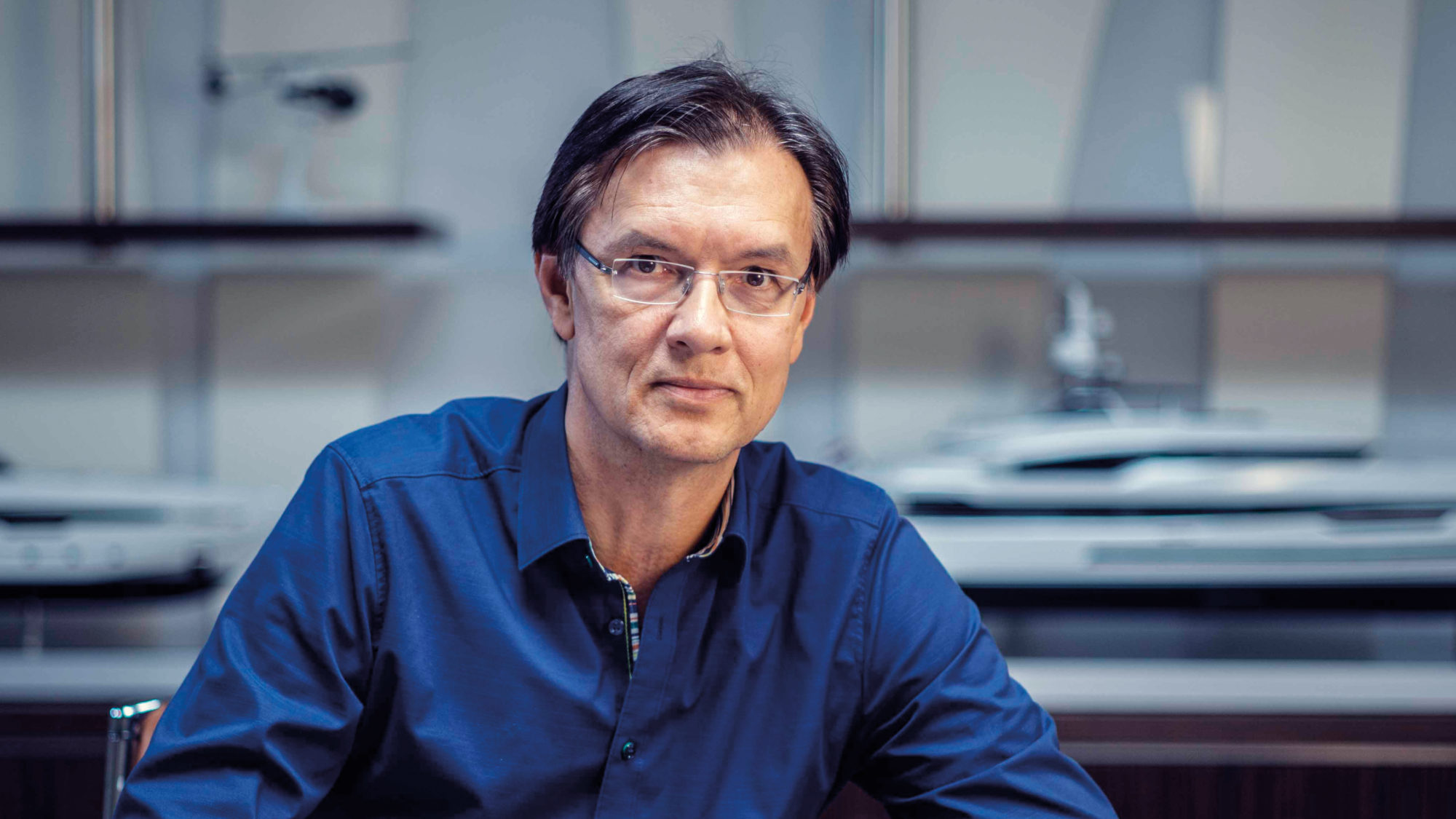 A man wearing glasses and a blue shirt seated in an office with scale models of boats in the background.