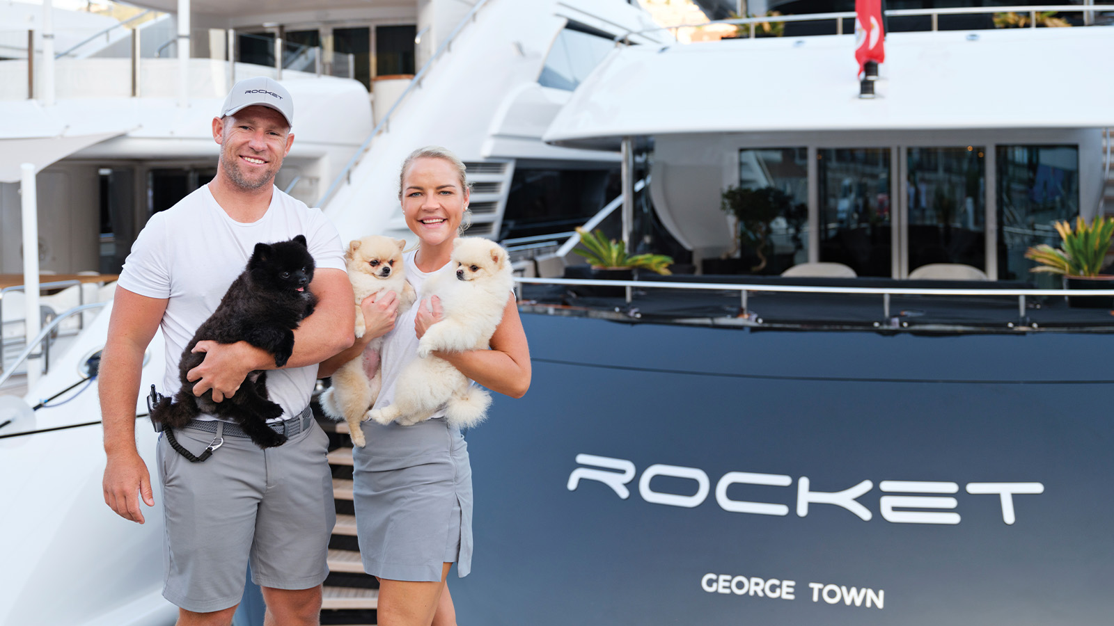 A man and woman standing in front of the yacht 'Rocket' holding three small dogs.