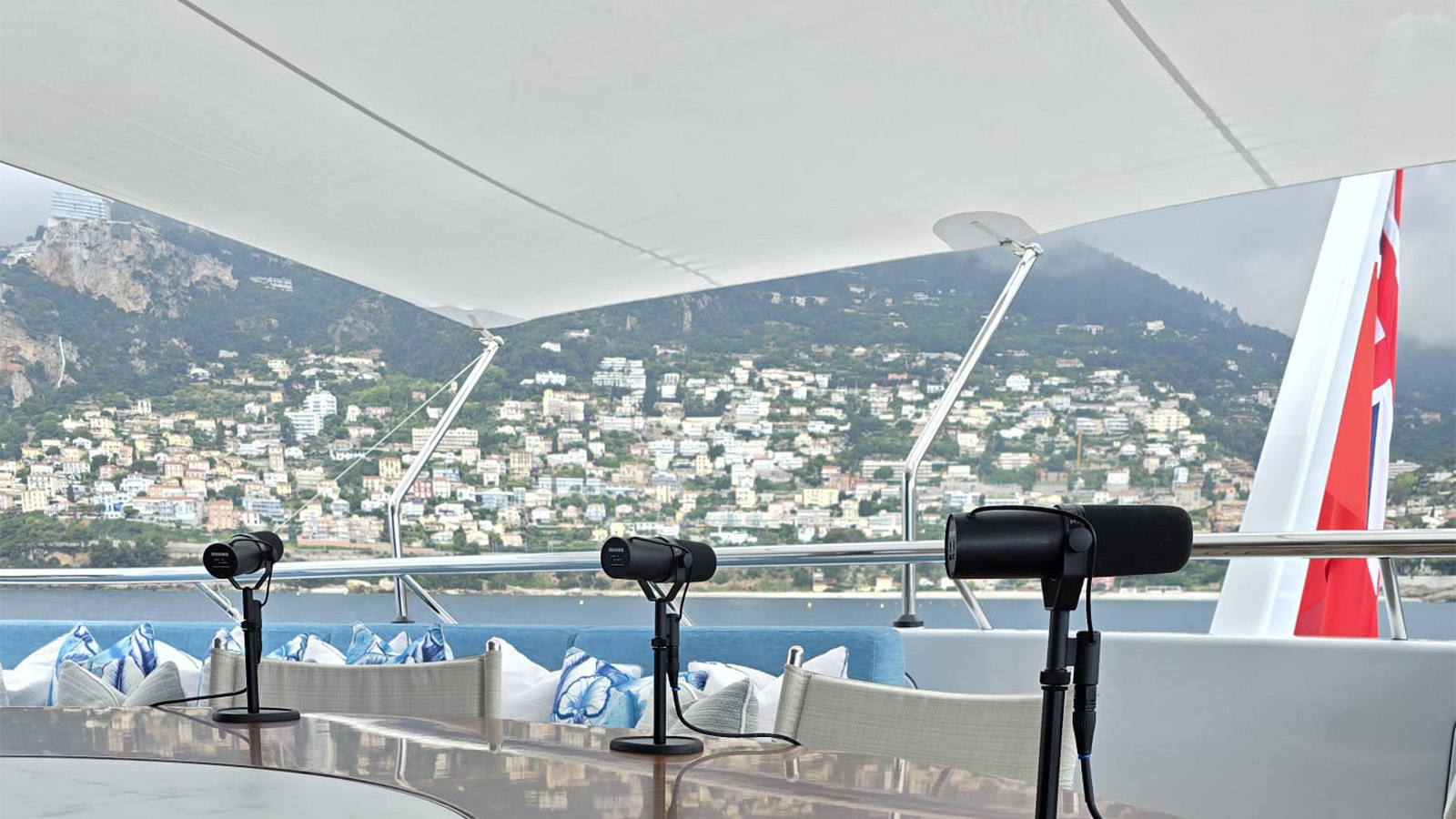 A modern outdoor setting on a boat, featuring a table with three microphones, surrounded by blue and white cushions. In the background, there is a coastal town with numerous buildings on a hillside and a red flag is partially visible on the right side.