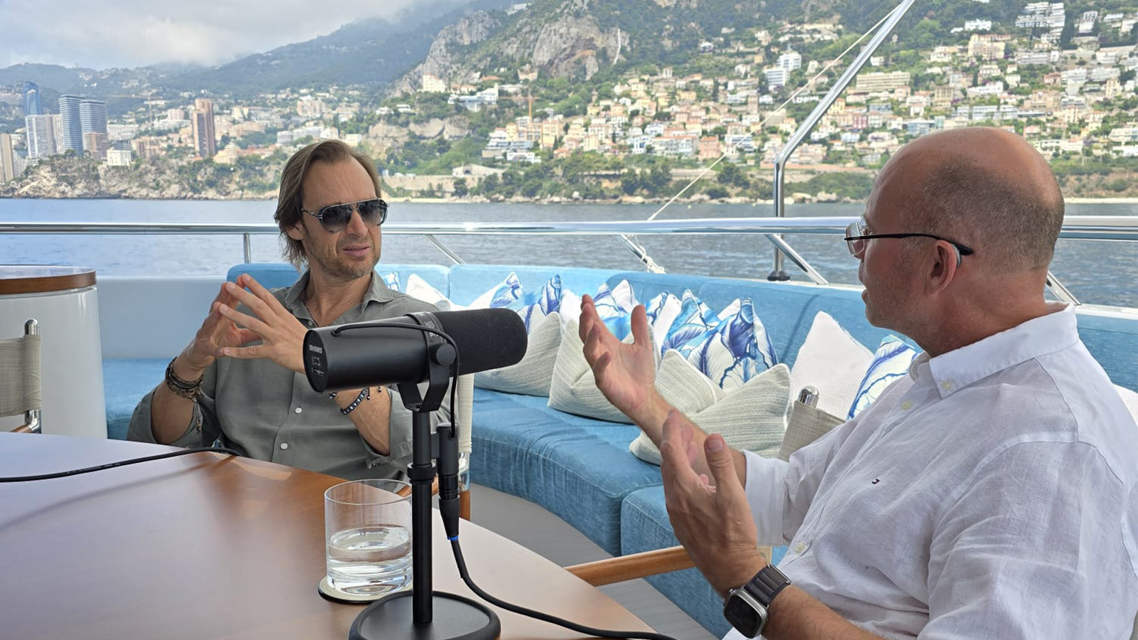 Two men engaged in conversation on a yacht, with a podcast microphone visible between them. The man on the left is wearing sunglasses and a gray shirt, while the man on the right is in a white shirt. The backdrop shows a scenic coastline with hills and buildings.