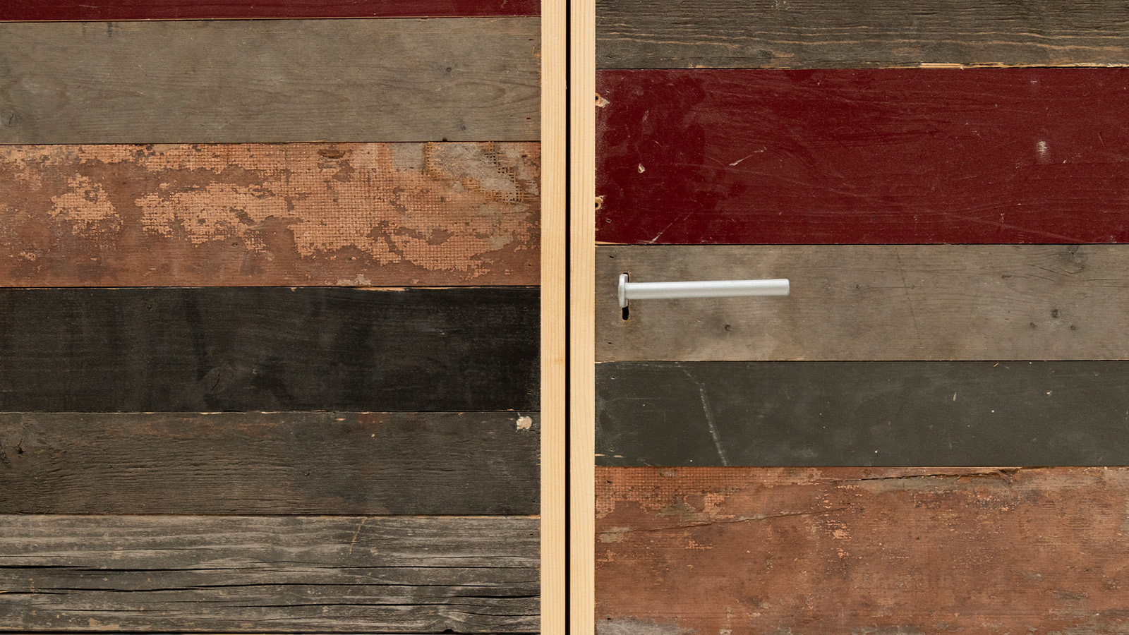 A wooden door made of horizontally aligned, reclaimed wood panels in various shades of red, brown, black, and gray, featuring a simple metal handle on the right side.