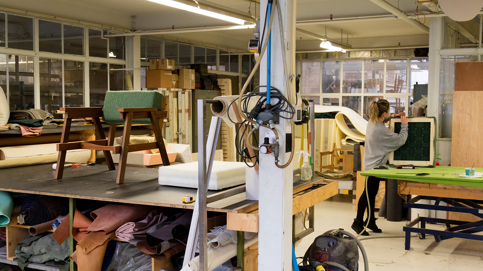 A person working in an upholstery workshop with various materials and tools spread across tables and shelves. The room is filled with fabrics, foam padding, and wooden furniture frames, indicative of a crafting environment.