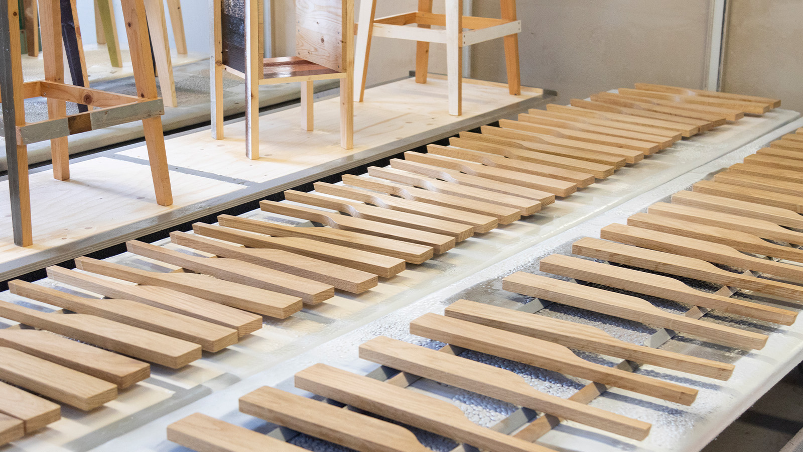 A workshop with neatly arranged wooden planks, likely for furniture construction, and in the background, several stools on display.