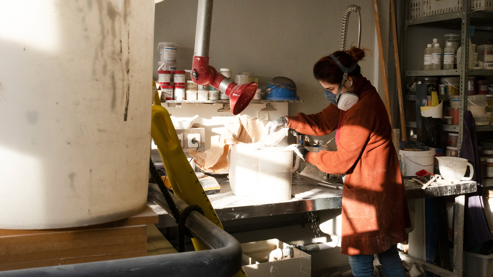A person wearing a mask and gloves, working with plaster in a workshop filled with supplies and tools.