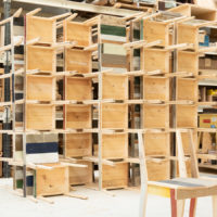 A workshop filled with wooden shelving units and various wooden materials organized on shelves, with chairs partially visible in the foreground.